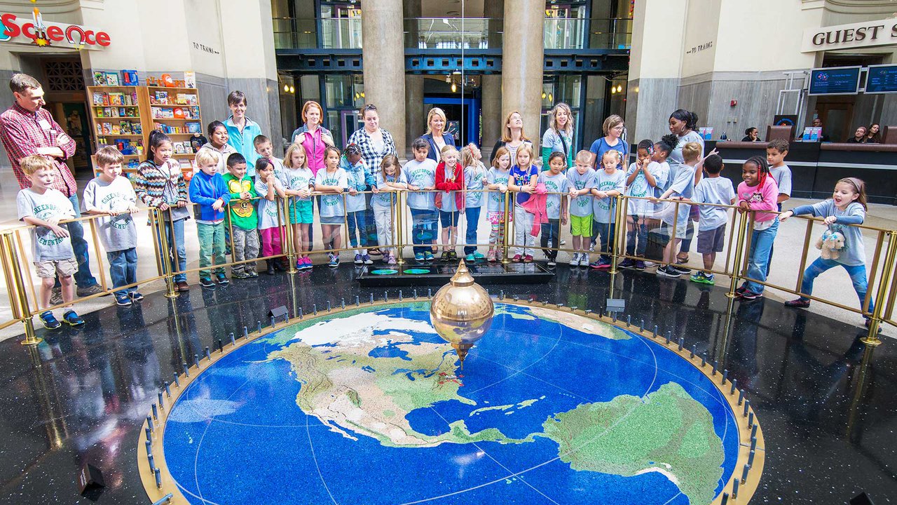 A photo of guests on a field trip at the Science Museum standing around the outside of the barrier around the pendulum.