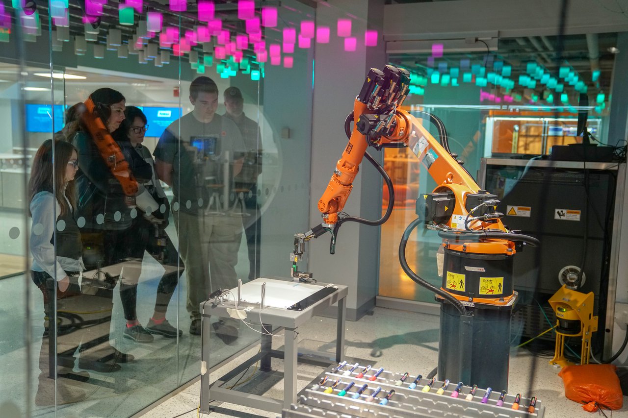 Group of five people watching a large orange machine through a glass window.