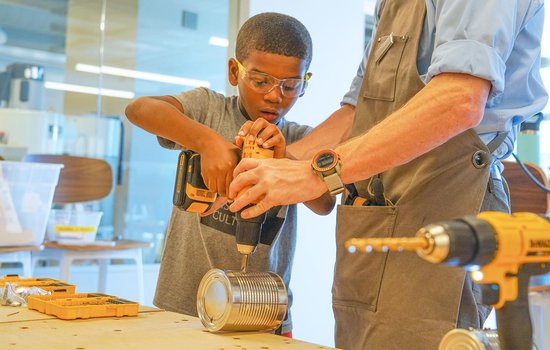 A young boy wearing safety goggles helping an adult man drill into a metal can.