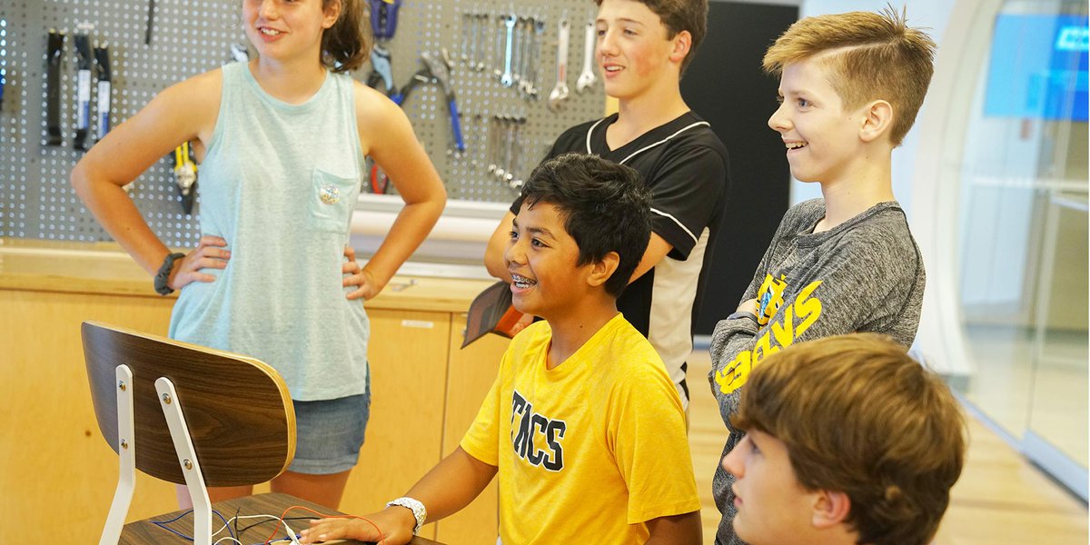Five children standing in front of a wall with tools hanging on it, smiling excitedly.