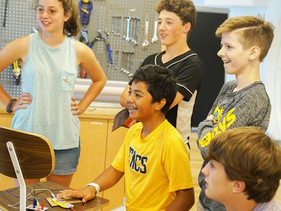 Five children standing in front of a wall with tools hanging on it, smiling excitedly.