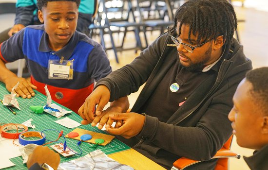 Three young men sitting together at a table working with different materials.
